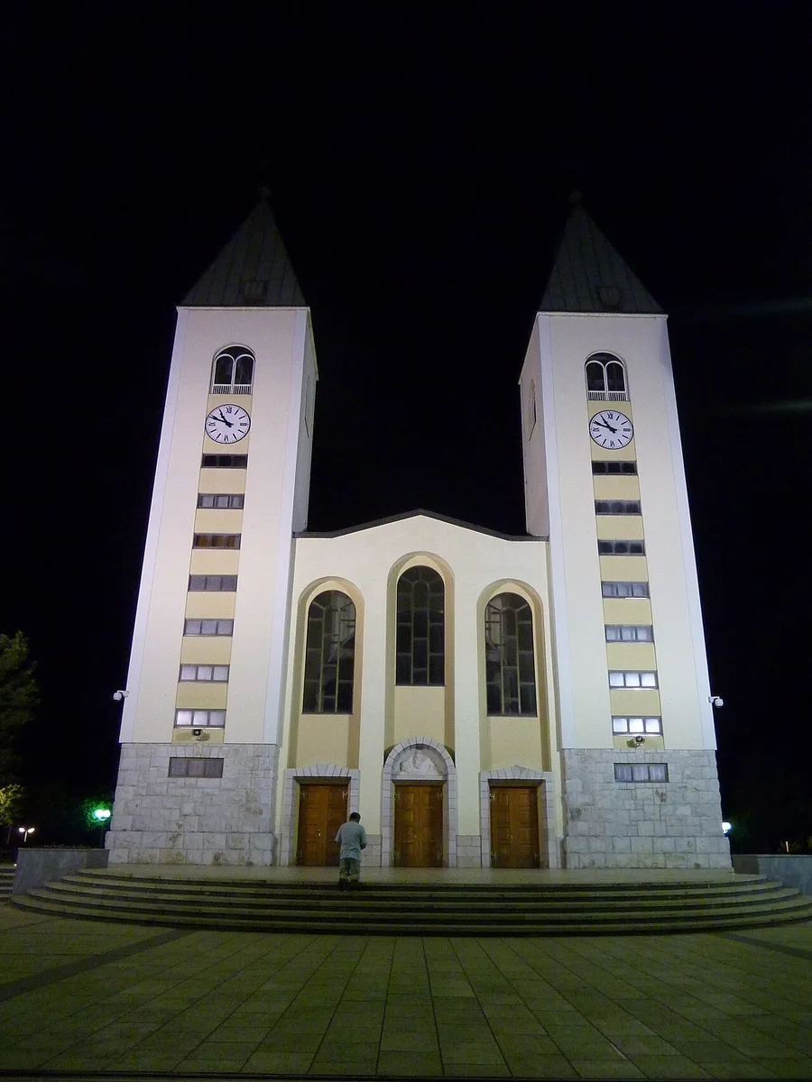 The twin bell towers of St James Church in Medjugorje floodlit at night, a lone figure at the doors