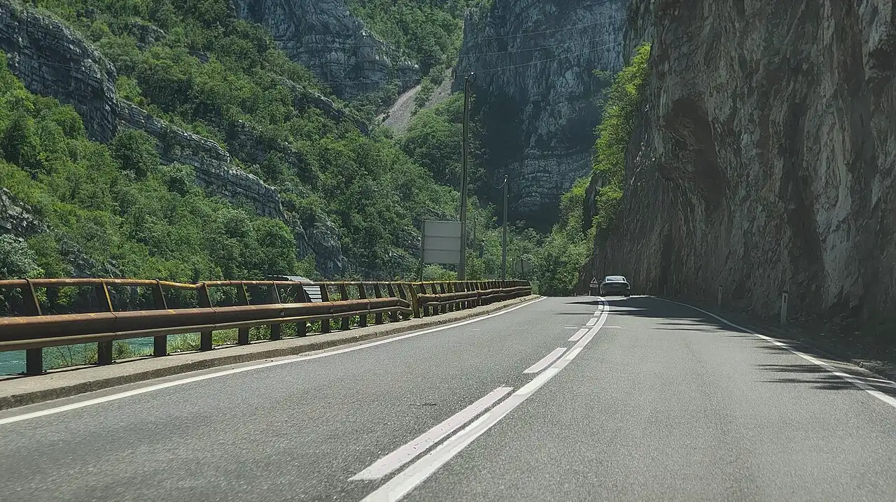 An open two-lane road curving through green hills in Bosnia and Herzegovina under a bright sky, a single car in the distance
