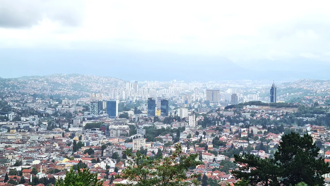 Panorama of Sarajevo filling its valley, minarets and red roofs below green hills, the destination of the route