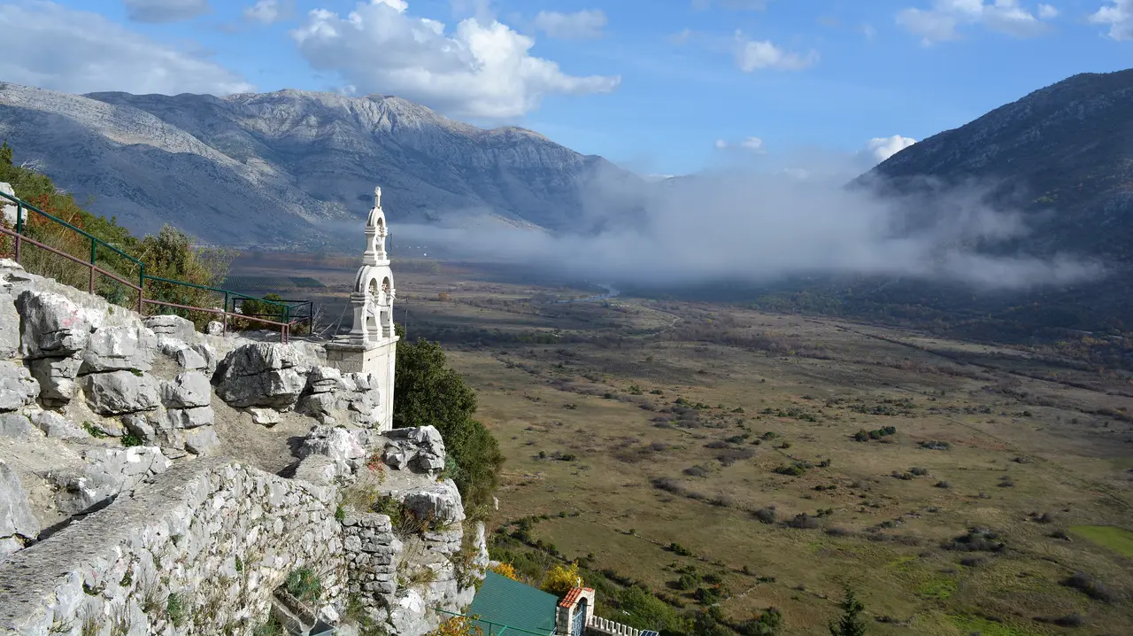 The white stone bell tower of Zavala monastery on a karst ledge above Popovo polje, low cloud lying along the valley floor