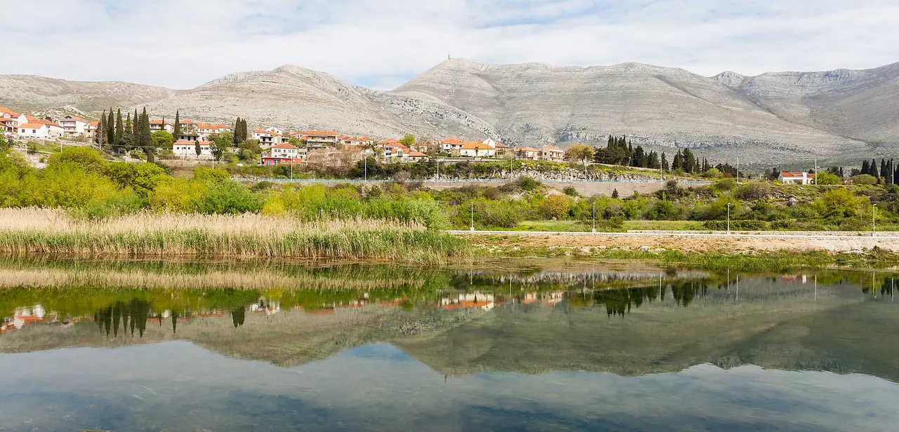 A village on the far bank of the Trebisnjica reflected in still water, with the grey ridge of Leotar rising behind