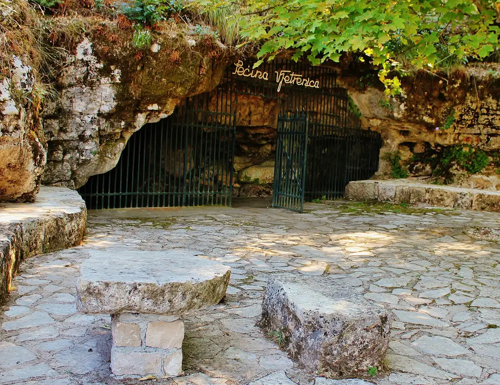 The barred gate of Vjetrenica cave under a mossy limestone overhang, stone tables and a paved forecourt in front