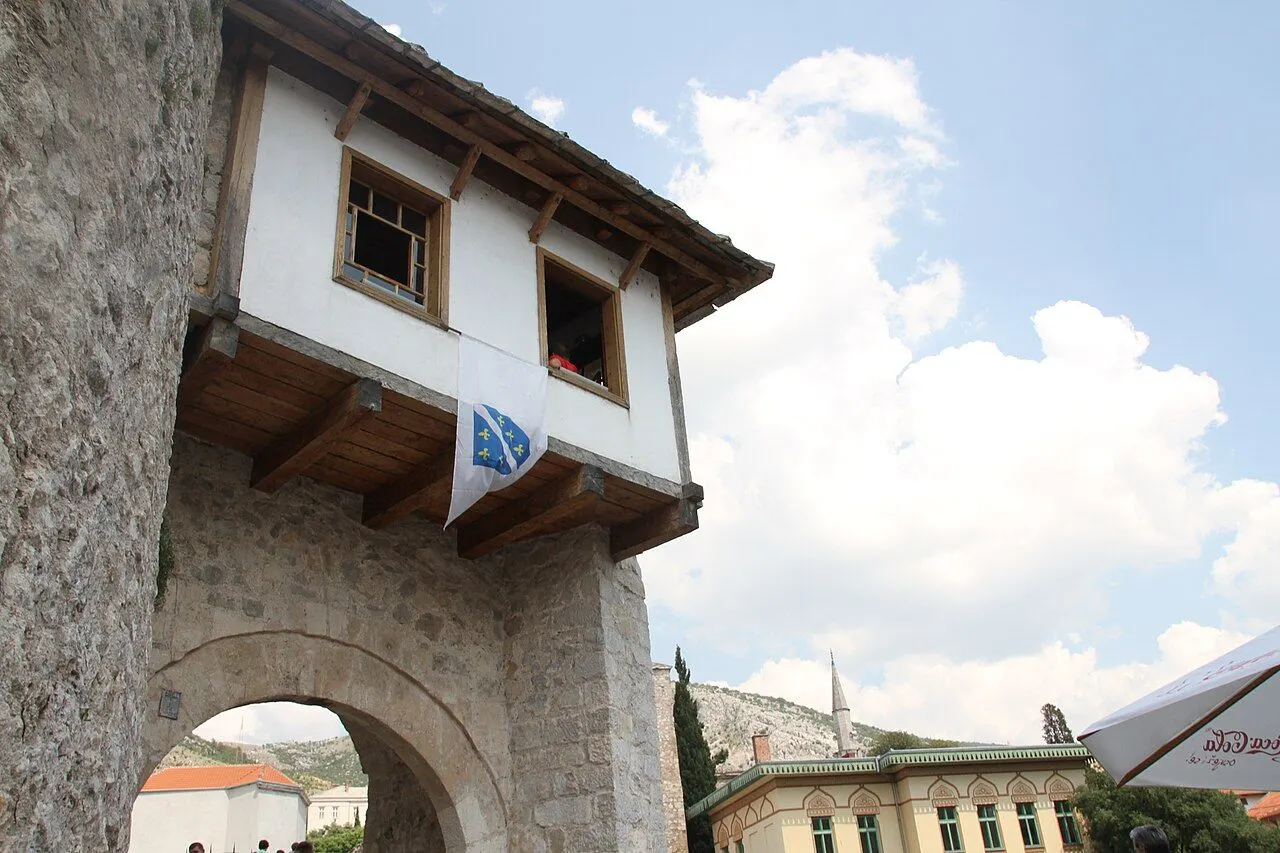 A traditional overhanging stone gatehouse with a Bosnian flag beneath its wooden eaves at the edge of Mostar old town