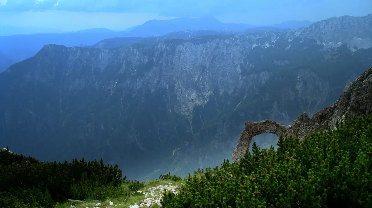 The Hajducka vrata rock arch on Cvrsnica mountain, with dwarf pine in the foreground and the hazy walls of the Diva Grabovica canyon behind