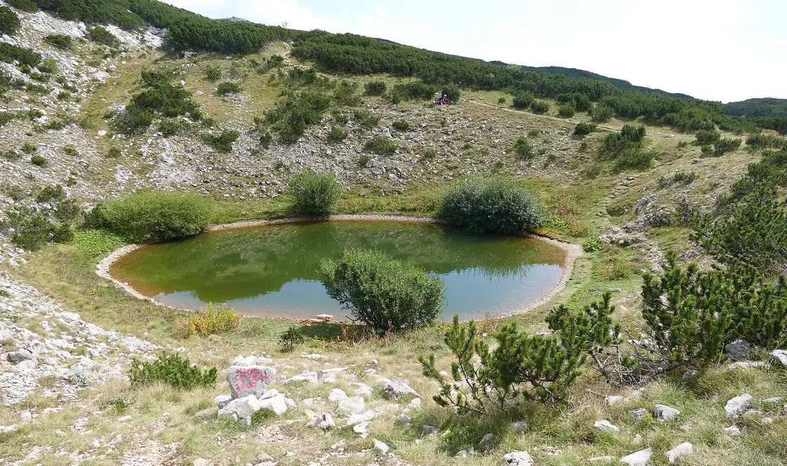 The small green Crvenjak tarn in a stony hollow on Cvrsnica, ringed by dwarf pine, with a painted trail waymark on a rock in front