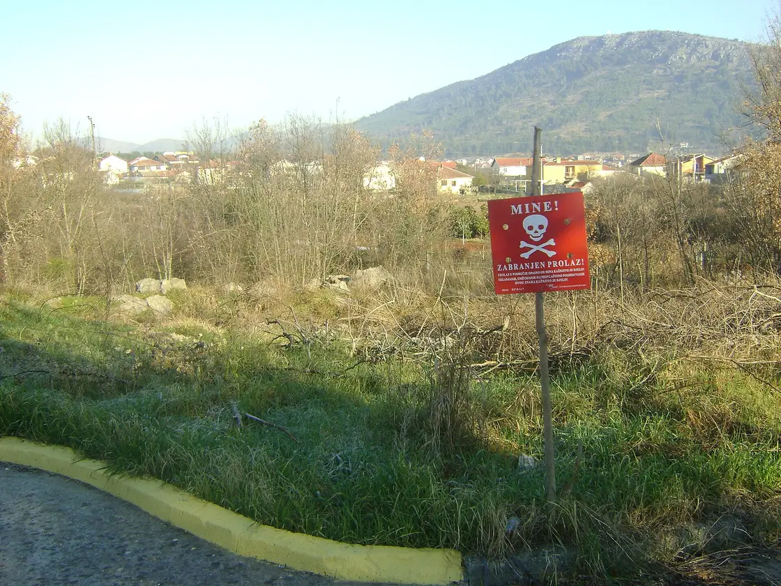 A red warning sign reading MINE! ZABRANJEN PROLAZ! on a post beside rough scrub at the edge of a road in Herzegovina, with houses and a hill behind