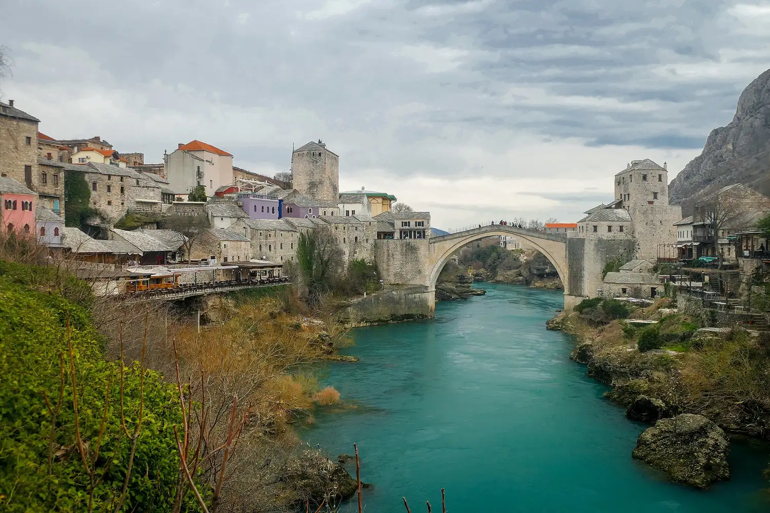 The single stone arch of the Old Bridge of Mostar over the green Neretva under a cloudy sky, stone towers on both banks