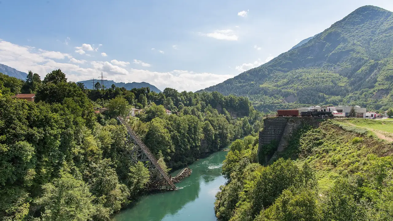 The ruined Narentabahn railway bridge over the green Neretva at Jablanica, steel lattice resting against the hillside