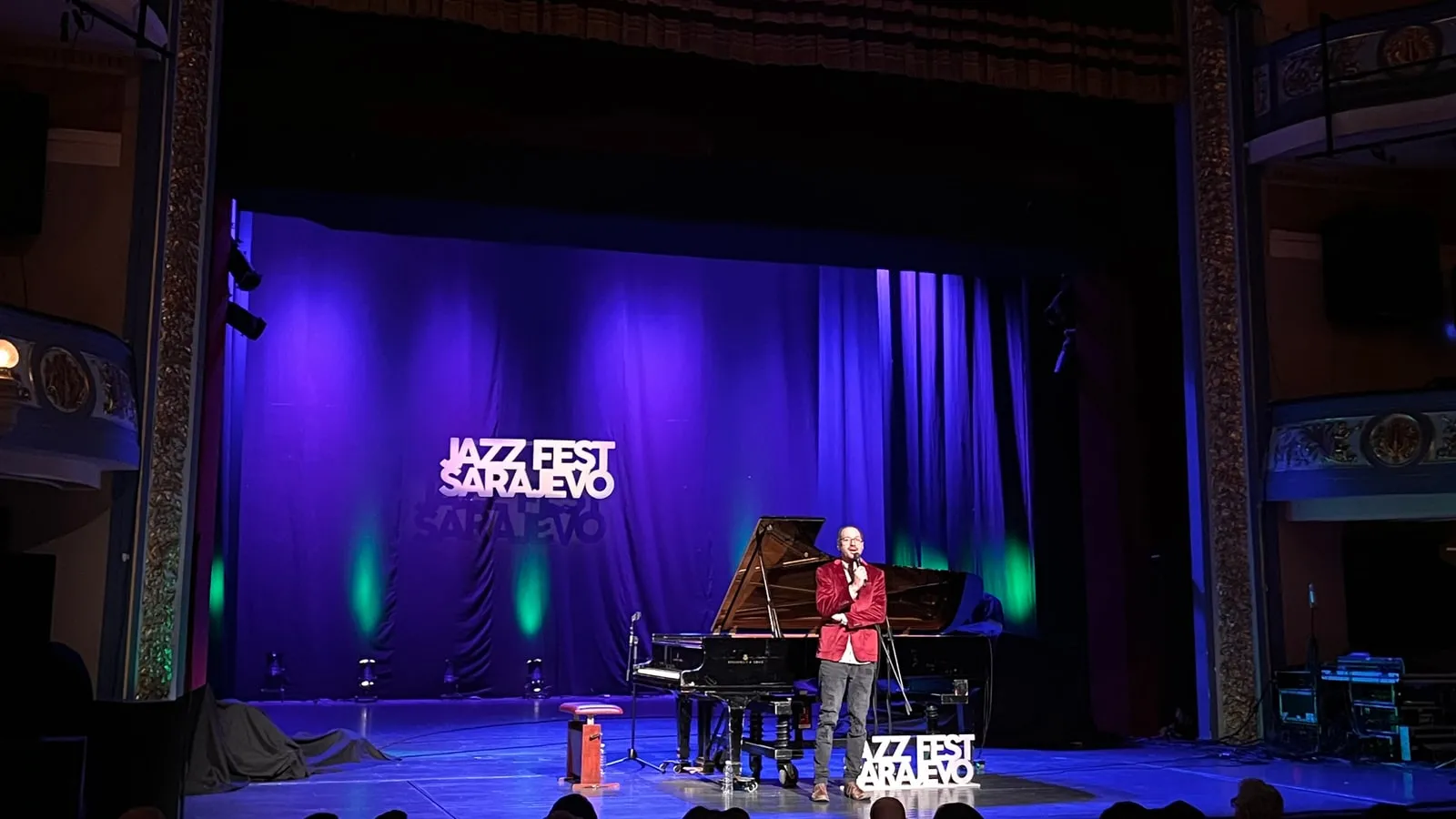 A presenter in a red jacket beside a grand piano on the blue-lit stage of the Sarajevo National Theatre, Jazz Fest Sarajevo logos on the backdrop and the stage floor