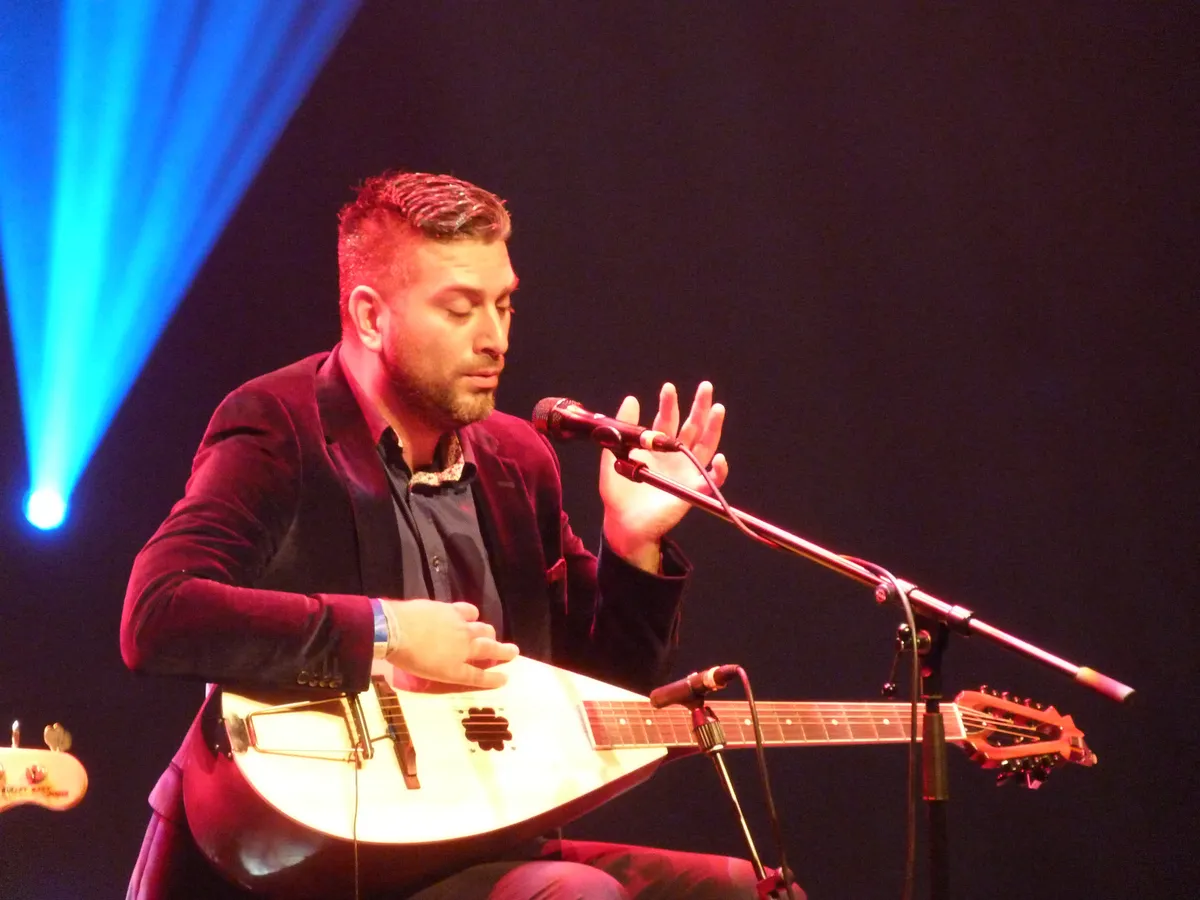 Damir Imamovic singing into a microphone on a dark stage, a long-necked white tambura across his lap and a blue spotlight behind him