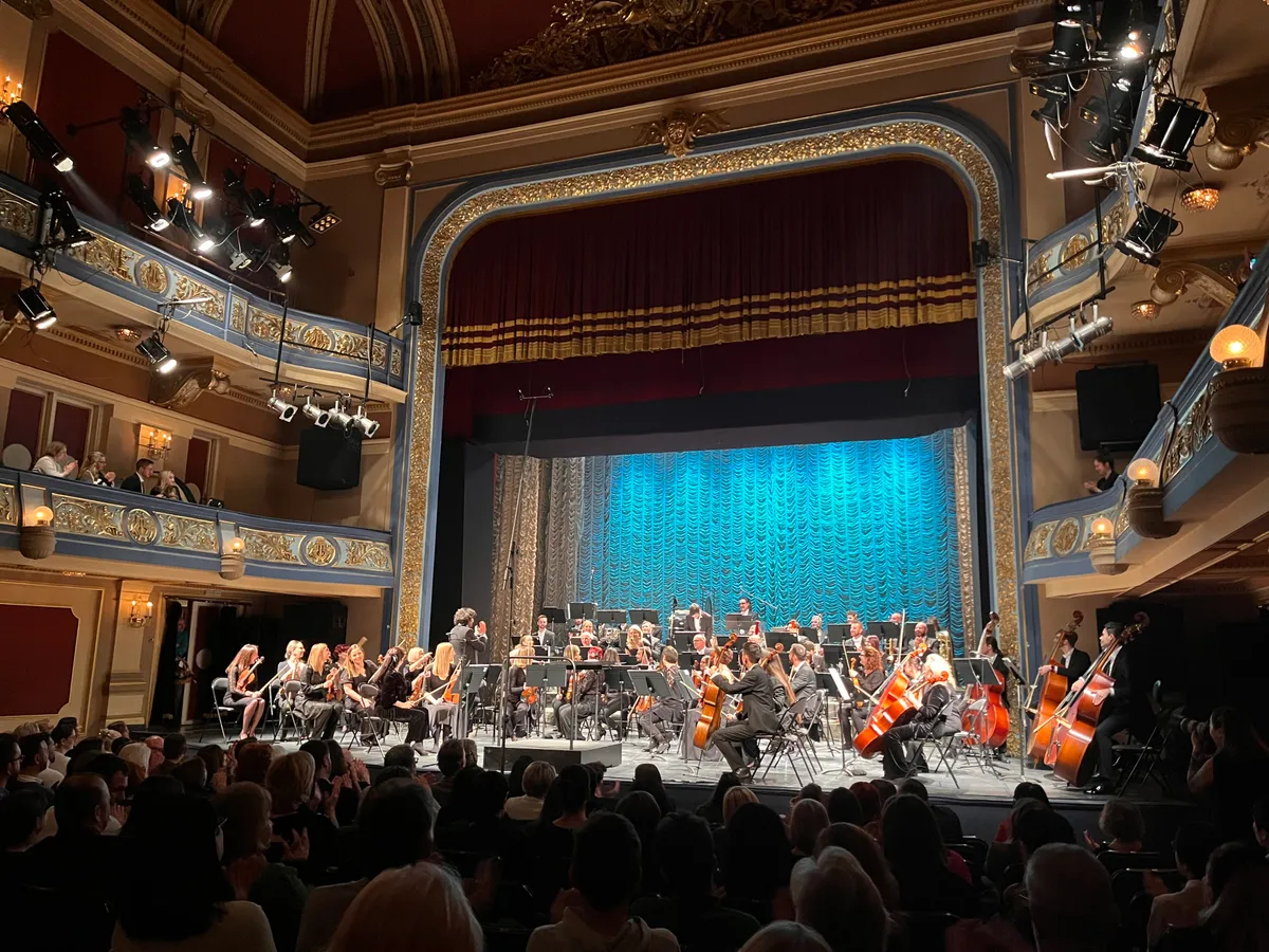 An orchestra on the stage of the Sarajevo National Theatre seen from the stalls, gilded boxes and balconies rising on both sides of the proscenium