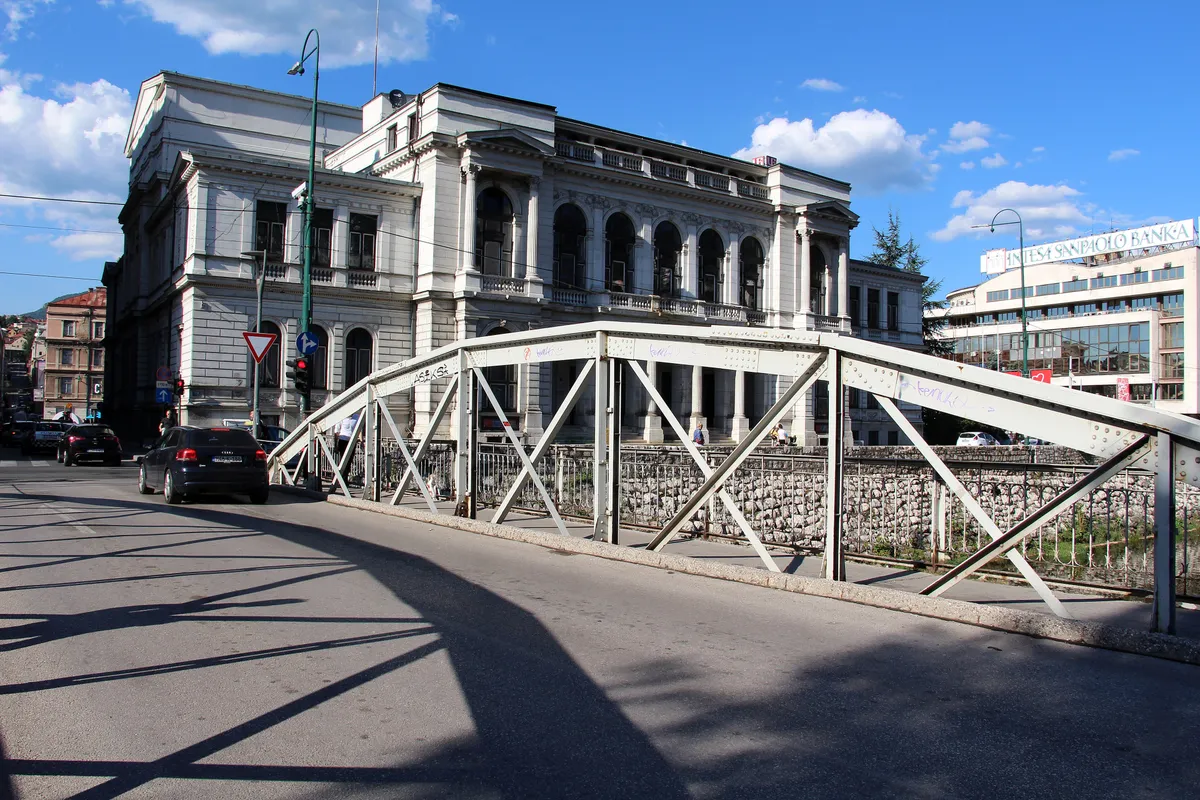 The white neo-Renaissance facade of the Sarajevo National Theatre seen across a riveted iron bridge over the Miljacka on a sunny day