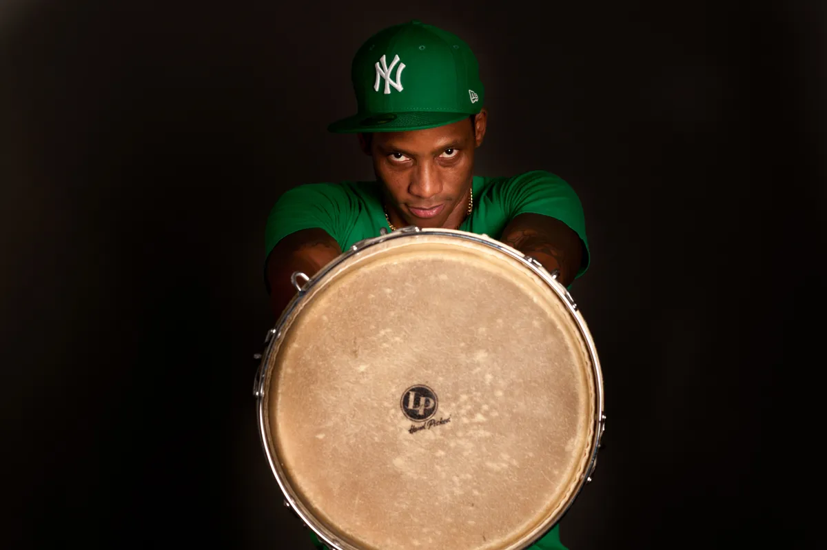 Cuban percussionist Pedrito Martinez in a green cap holding a conga drum head-on towards the camera against a black background