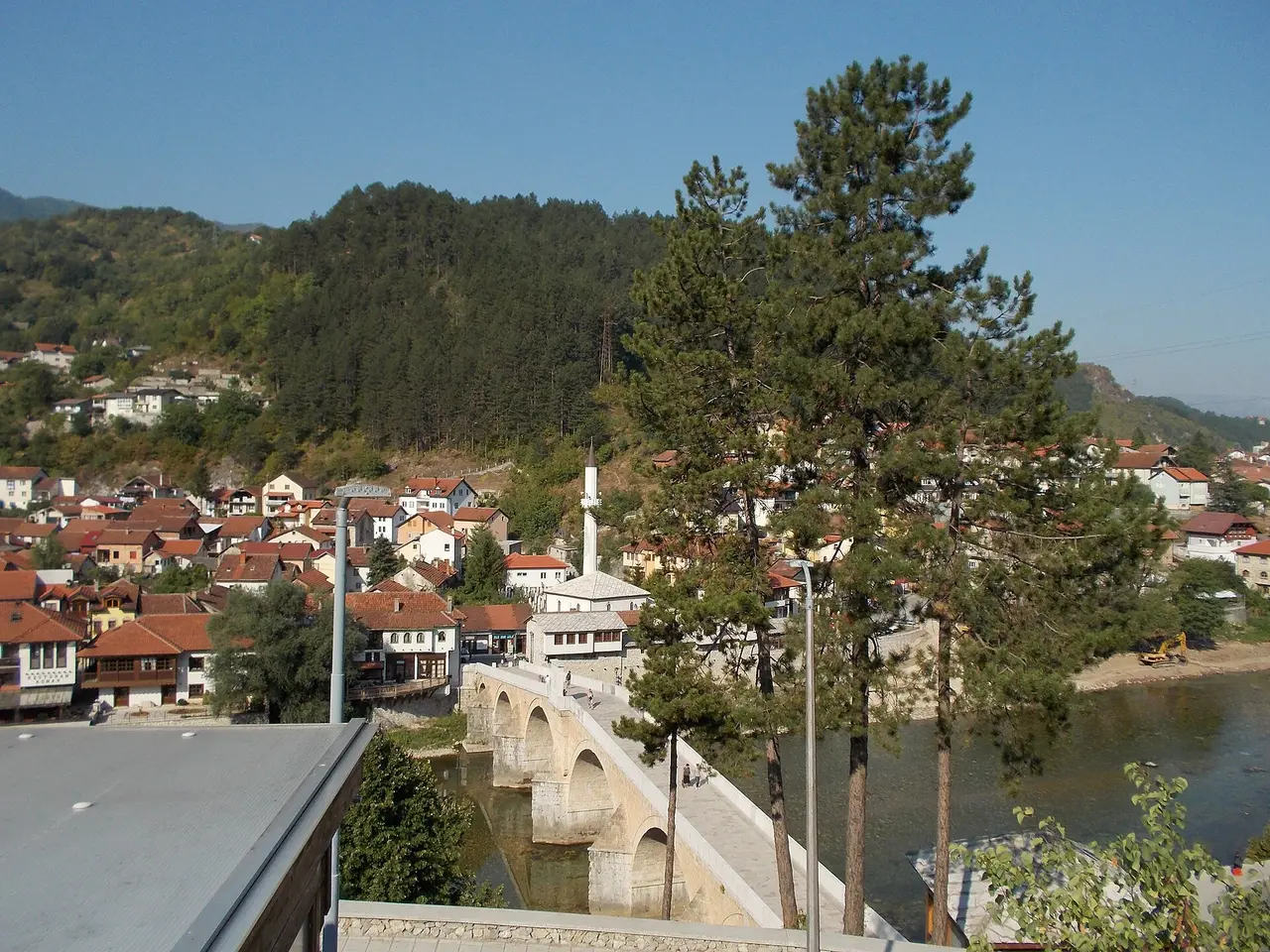 The six-arched Ottoman stone bridge of Konjic across the green Neretva, the town and a minaret rising on the far bank under wooded hills