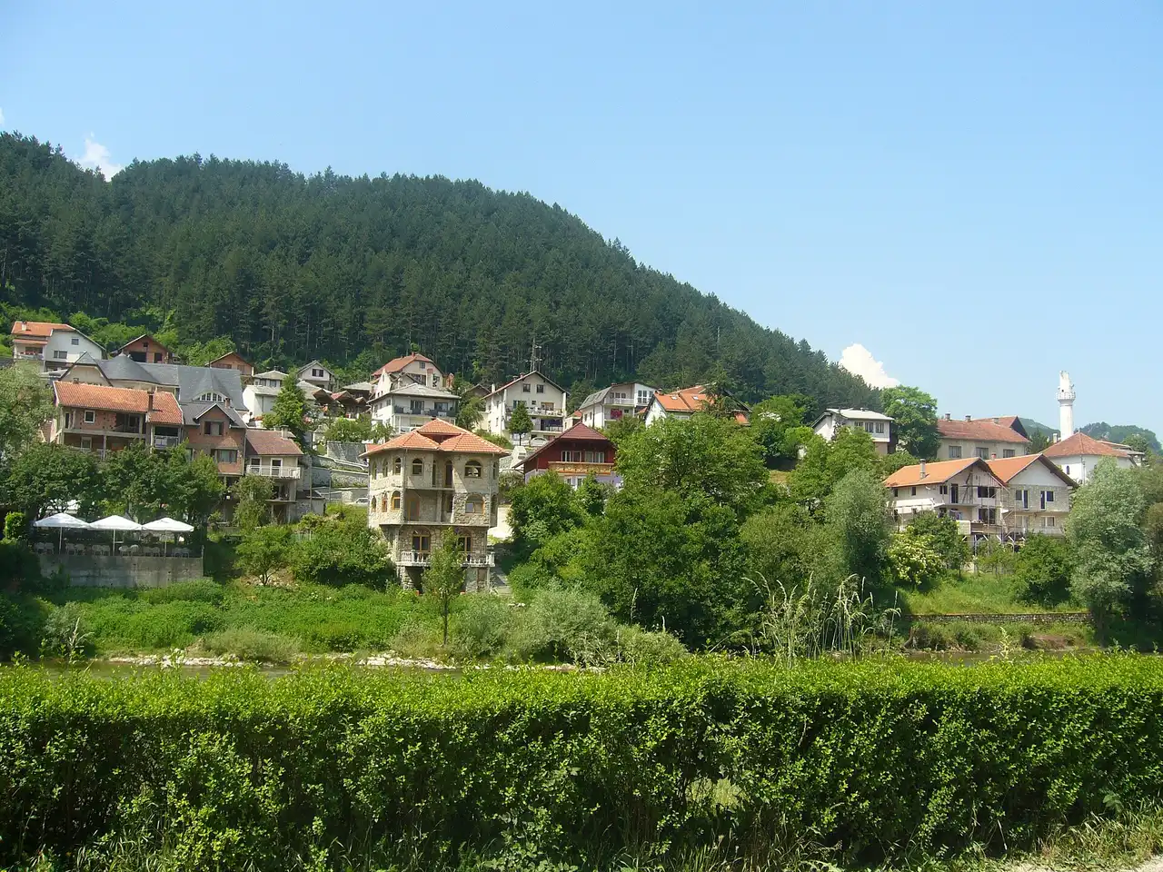 A hillside neighbourhood of Konjic with houses and gardens climbing the green slope, a minaret among the roofs