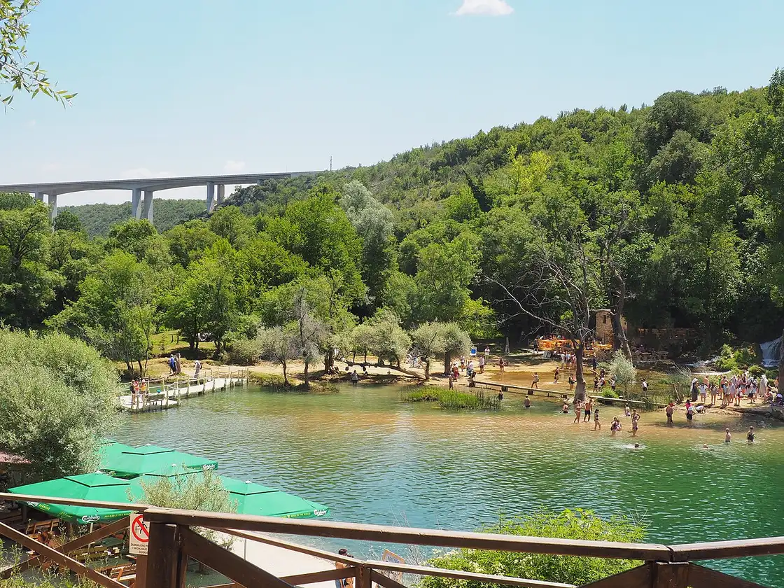The Kravice basin from a restaurant terrace with parasols, a wooden footbridge across the pool, swimmers and a motorway viaduct behind