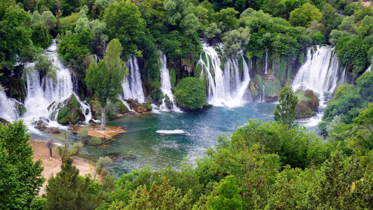 The broad horseshoe of Kravice Waterfalls seen from above, dozens of white cascades ringing a wide green pool in a forested basin