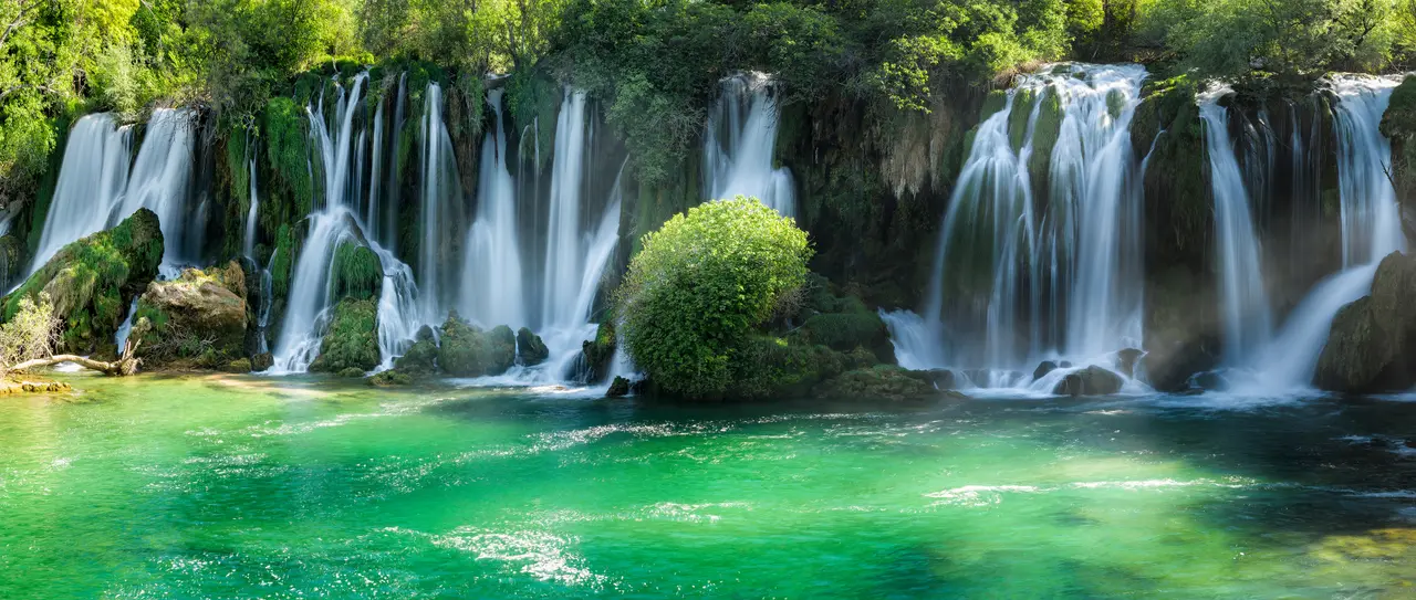 The tufa curtain of Kravice up close, ribbons of water pouring over moss-green travertine into a turquoise pool