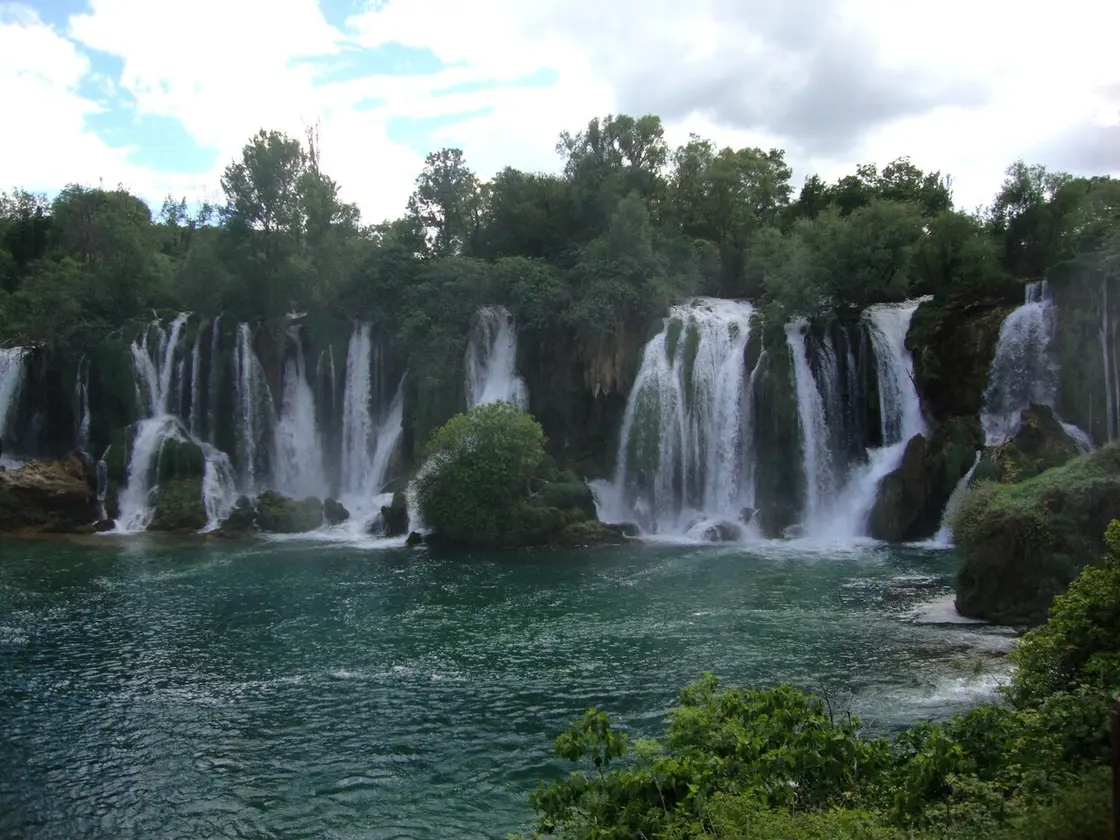Kravice waterfalls in fuller flow seen head-on from across the pool, the whole ledge running white under a cloudy sky