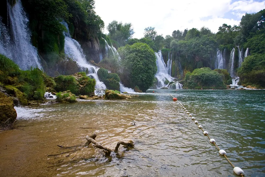 The swimming area at Kravice, a rope-and-buoy line marking the shallow zone off a sandy shore with the cascades behind