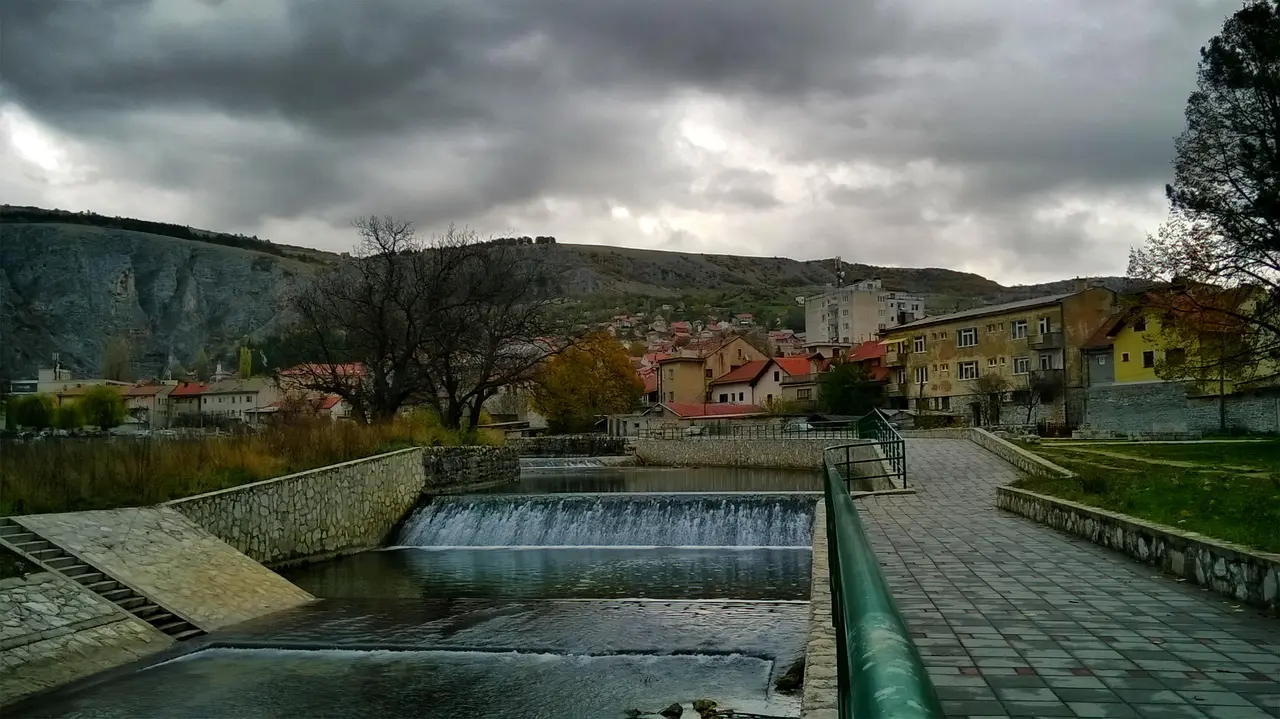 The small Bistrica river running through central Livno past stone embankments and pastel-coloured houses under a cloudy sky