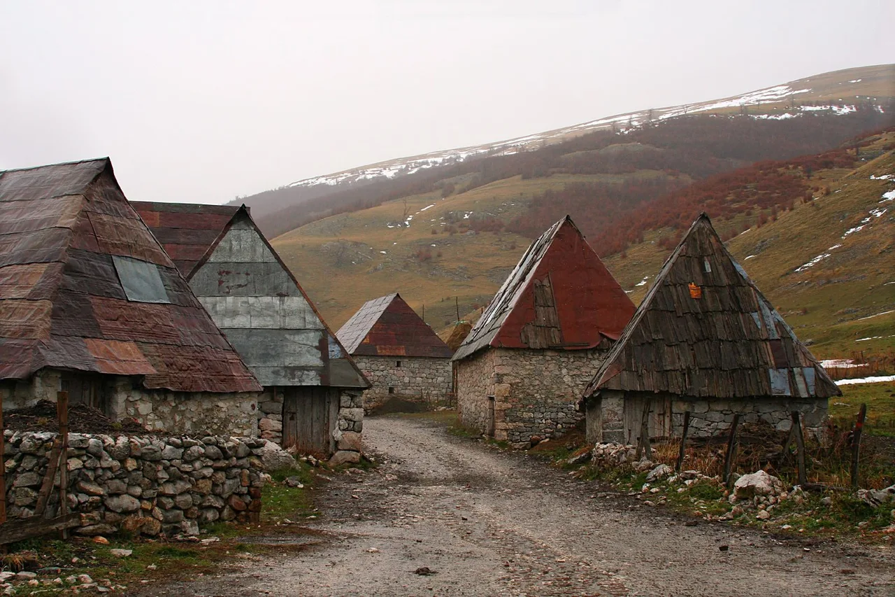 A row of stone houses with steep grey and red wooden-shingle roofs along a track in Lukomir, bare green mountains behind and no trees in sight