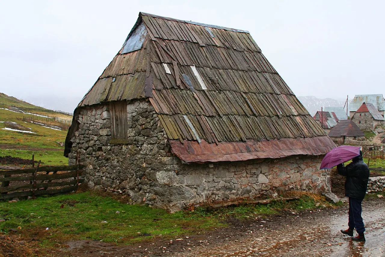 A single old stone house in Lukomir with a steep weathered wooden-shingle roof, a person sheltering under an umbrella beside its wall in the rain