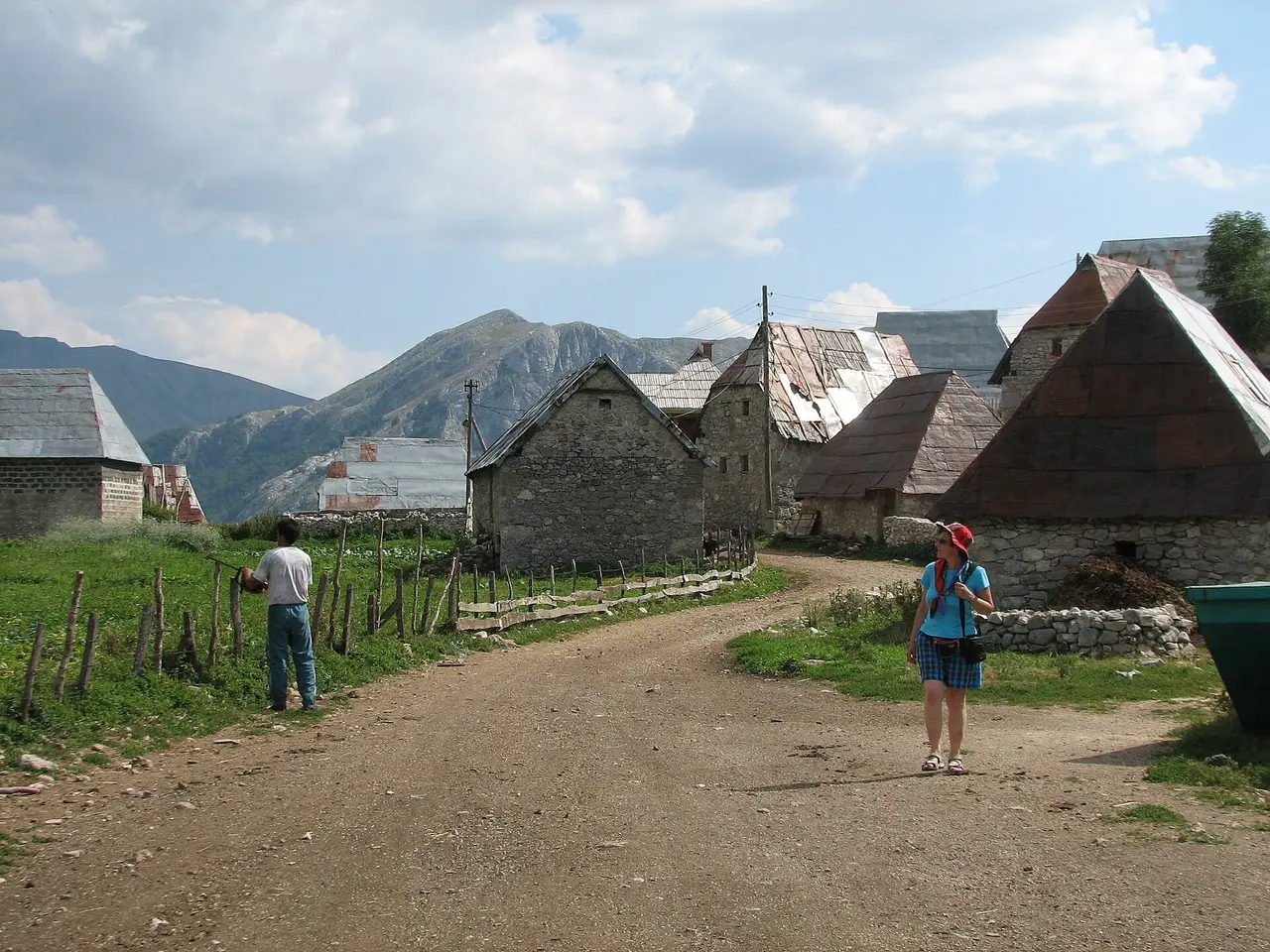 The single track through Lukomir lined with stone houses under shingle roofs, a resident and a visitor standing in the lane, high bare mountains behind