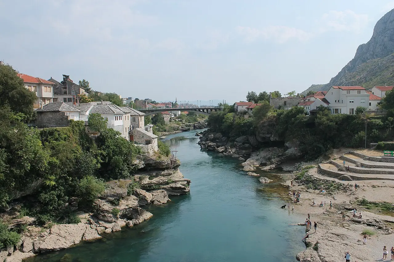 The turquoise Neretva river running through the old part of Mostar between stone houses and a rocky riverbank, with a modern bridge behind