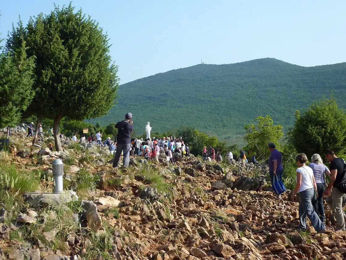 Pilgrims climbing the rocky karst path of Apparition Hill (Podbrdo) toward a white statue of the Virgin, a green ridge behind
