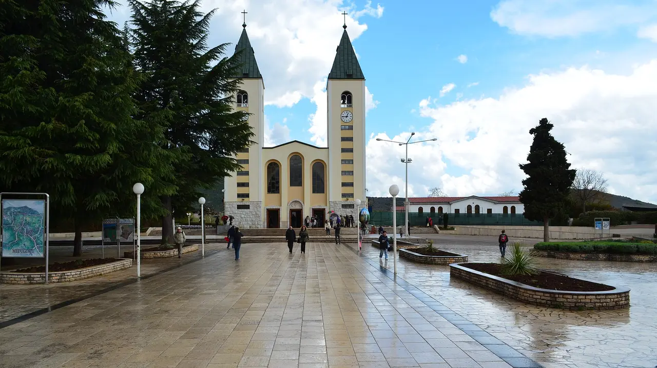 The twin bell towers of St James church in Medjugorje above a wide paved plaza with pilgrims walking