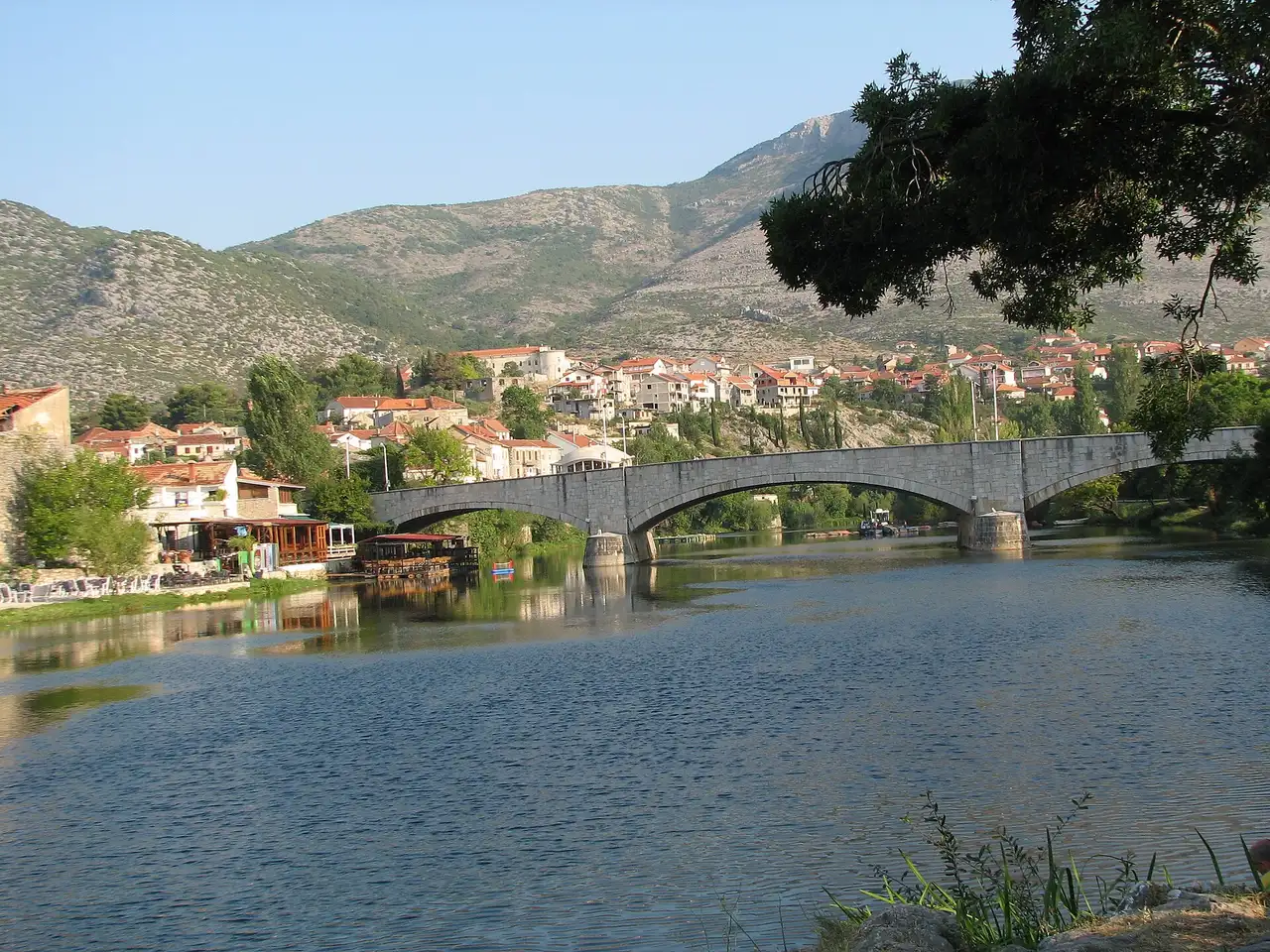 A long multi-arched stone bridge over the Trebisnjica river at Trebinje, with the town and bare mountains beyond
