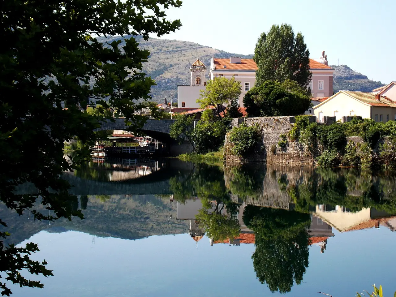 The old town of Trebinje reflected in the still Trebisnjica river, with a stone bridge and hills behind, the natural stop on the road to Kotor