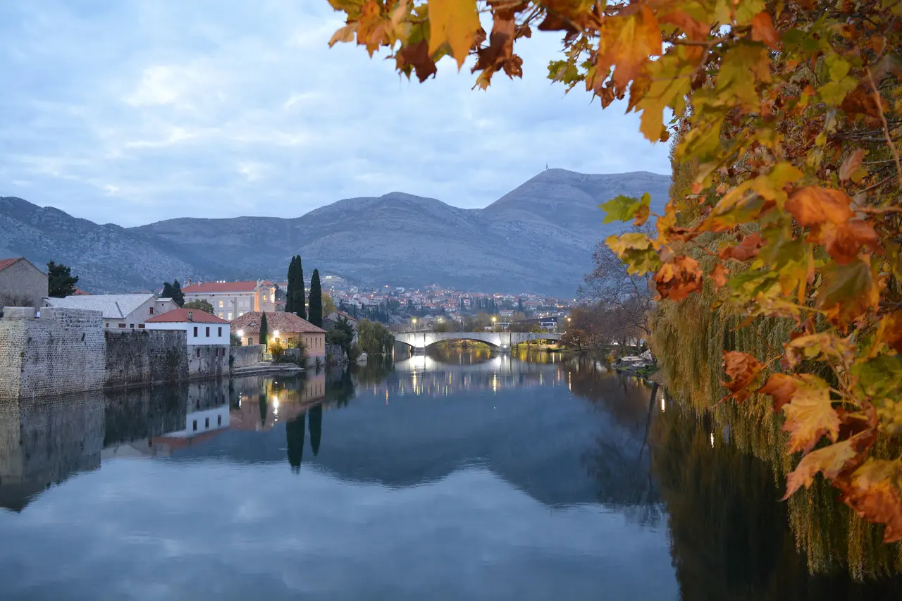 The old town of Trebinje beside the Trebišnjica river at dusk with bare autumn branches in the foreground
