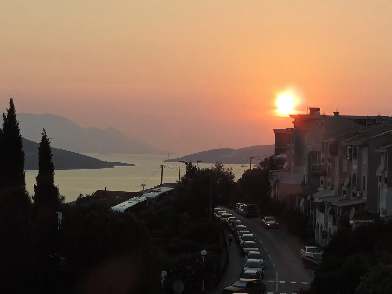 Sunset over Neum bay with the coast road lined with parked cars descending toward the water