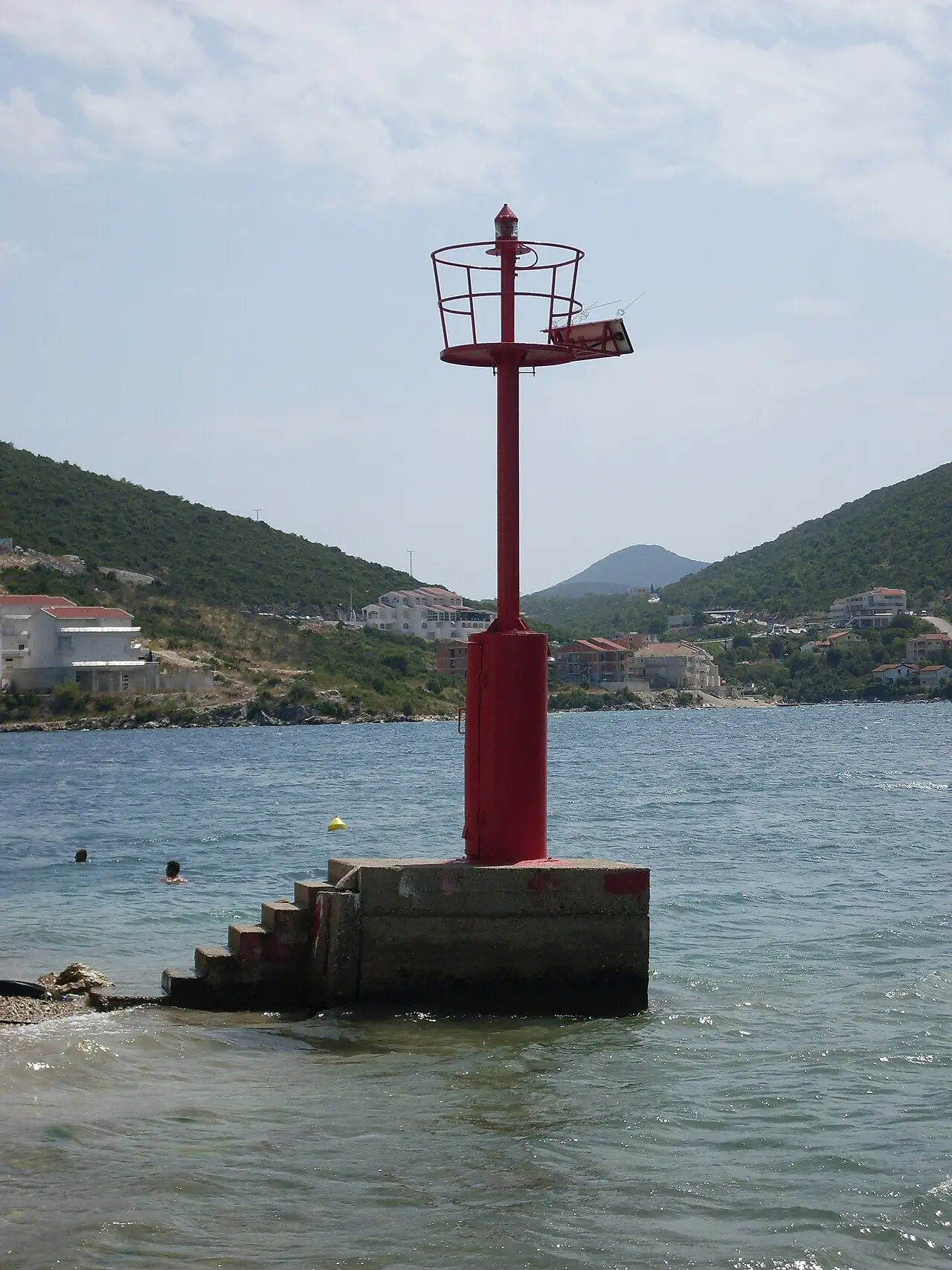 A red harbour beacon on a concrete base at the edge of the Neum shore with people swimming nearby and the town on the hillside behind