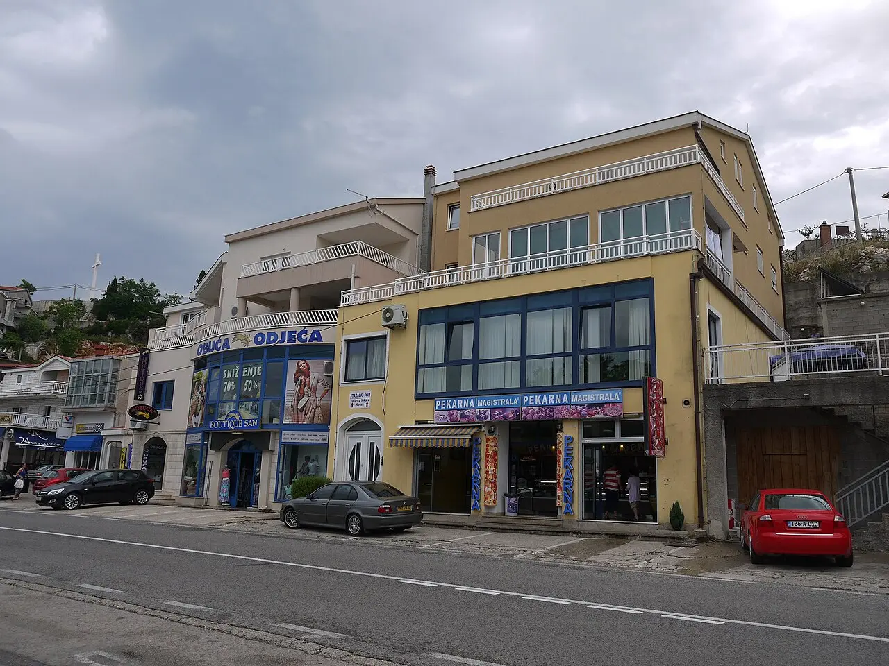 An everyday street in Neum with a bakery, a clothing shop and parked cars below houses climbing the hillside under a cloudy sky