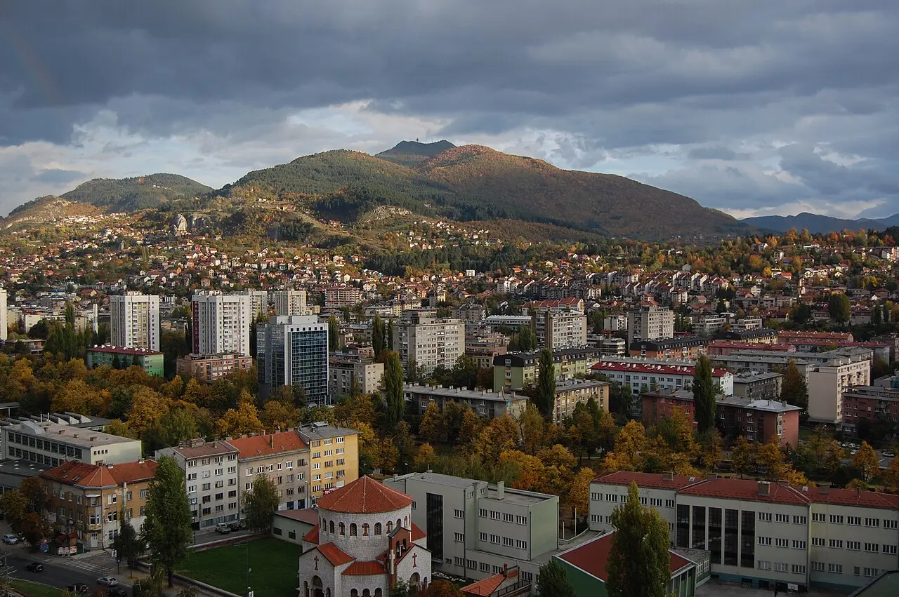 Autumn view over Sarajevo: pale apartment blocks and red roofs filling the valley below a long forested mountain ridge under a grey sky