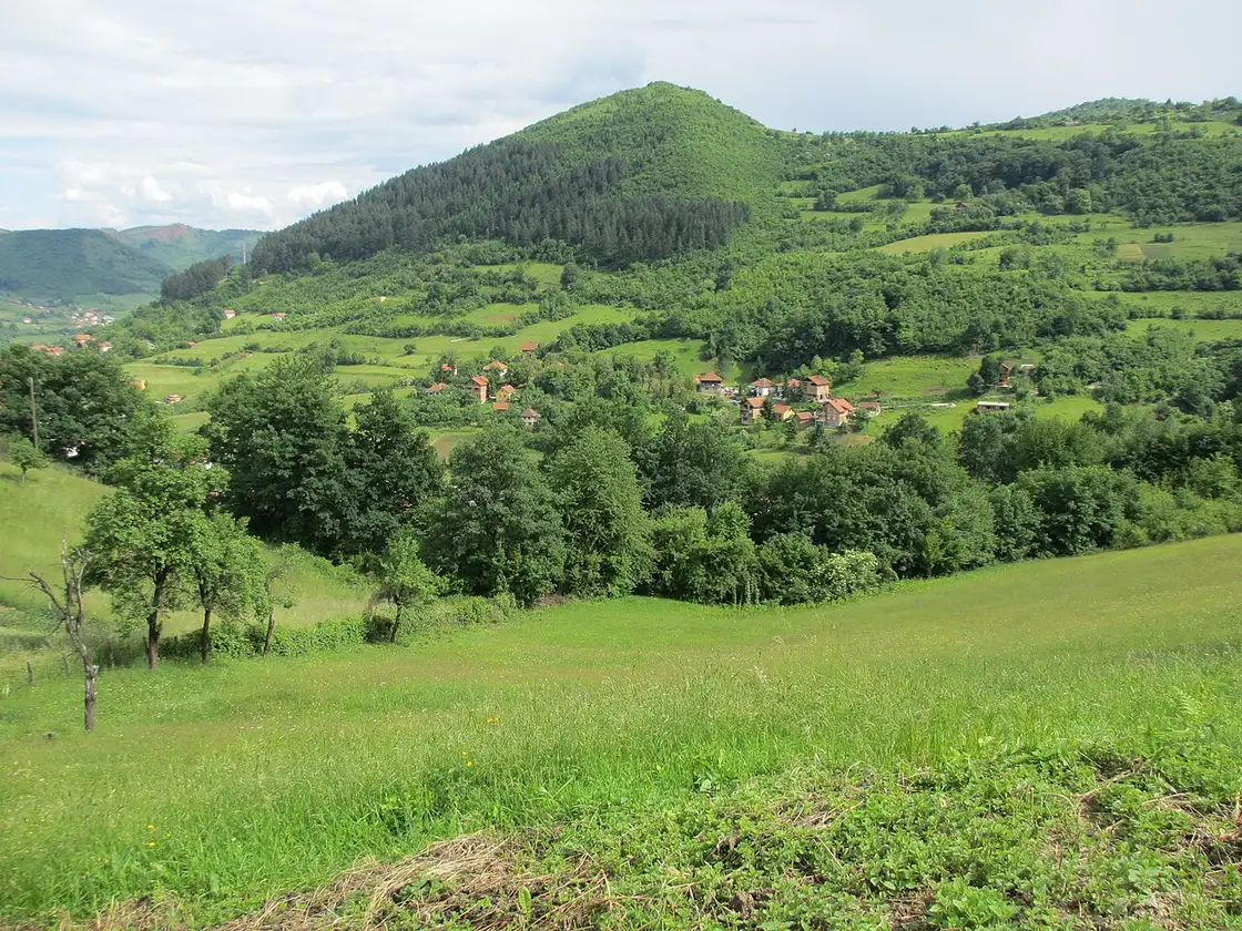 The steep, forested, near-symmetrical cone of Visocica hill rising above a green valley of scattered houses near Visoko, the Pyramid of the Sun