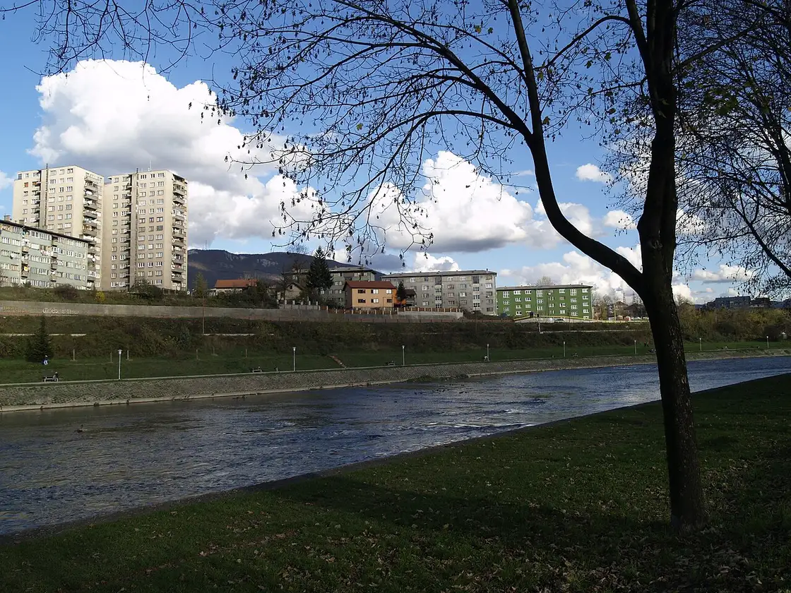 Socialist-era apartment towers lining the far bank of the Bosna river in Zenica, low green mountains behind under a blue autumn sky