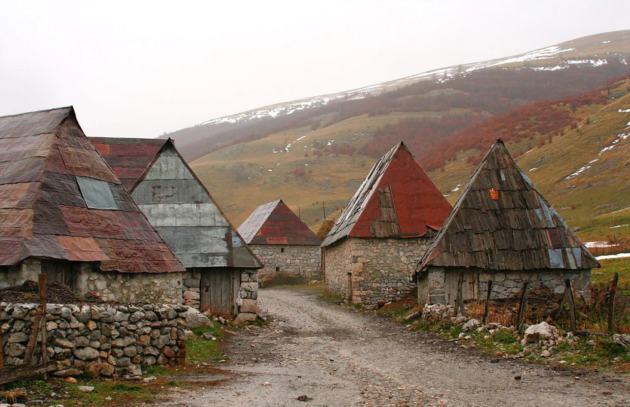 Stone houses with steep wooden-shingle roofs along a track in Lukomir village on a bare green ridge high on Bjelašnica