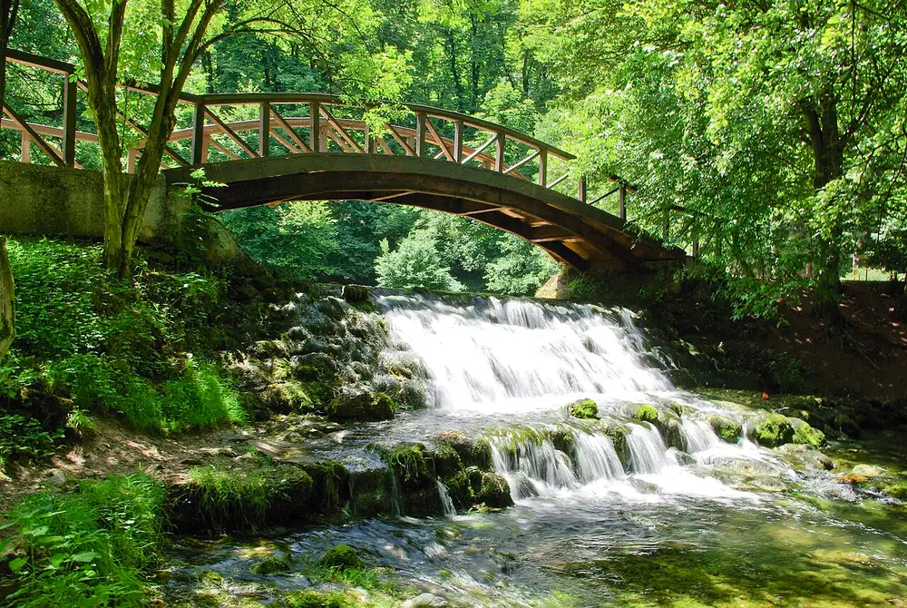 A small wooden footbridge over a clear, still channel at the Vrelo Bosne springs, green trees reflected in the water