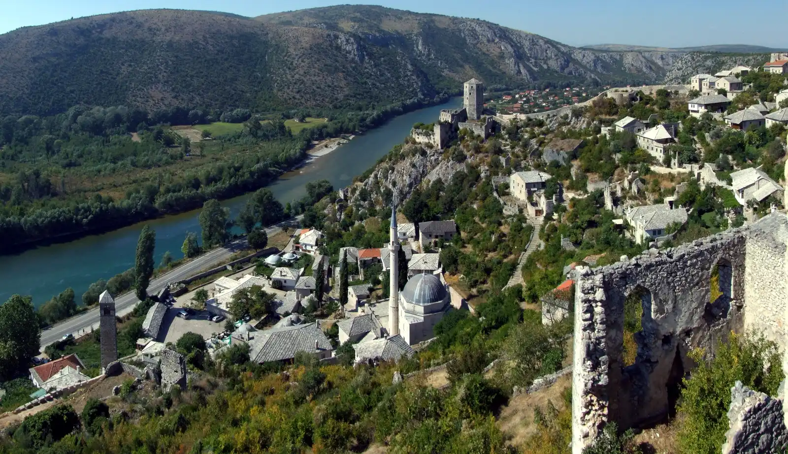 The Ottoman town of Počitelj climbing a limestone cliff above a bend in the green Neretva, the silo-shaped Kula tower, mosque dome and clock tower among stone houses
