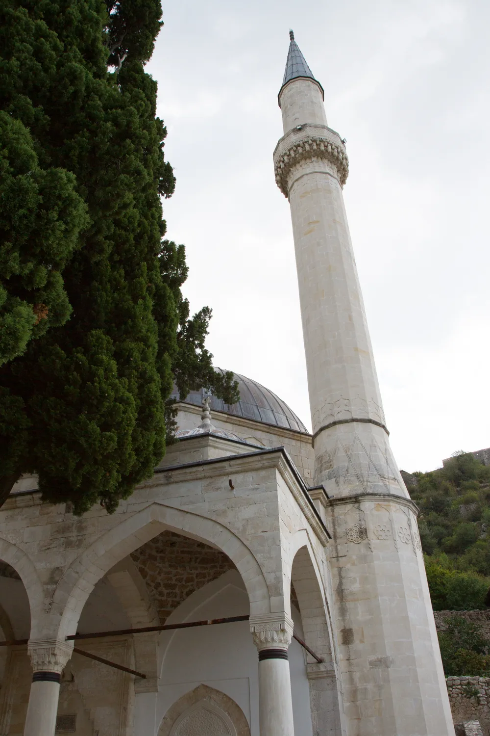 The domed Šišman Ibrahim-Pasha (Hajji Alija) mosque in Počitelj with its slender stone minaret and arched portico beneath a cypress tree