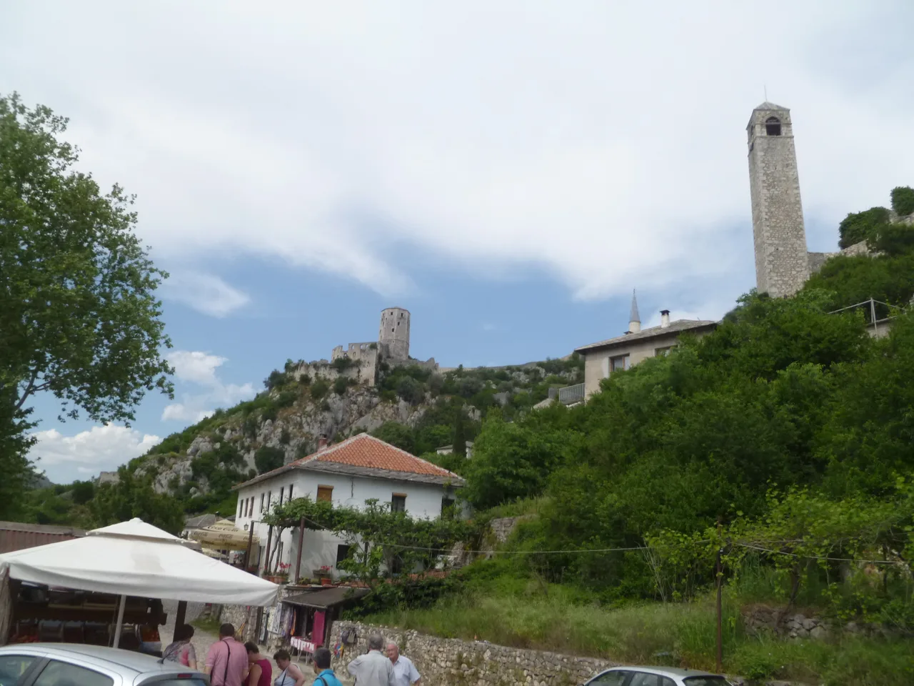 Stone houses, a souvenir stall with a parasol and the fortress towers of Počitelj seen from the roadside at the foot of the town