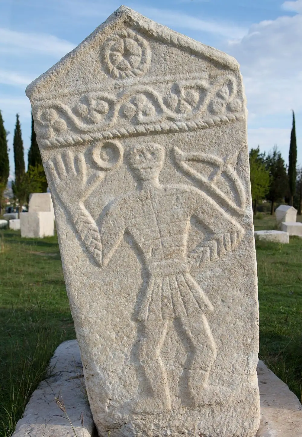 A tall stecak at Radimlja carved in relief with a standing male figure raising an oversized right hand, a rosette on the gable and a vine border above