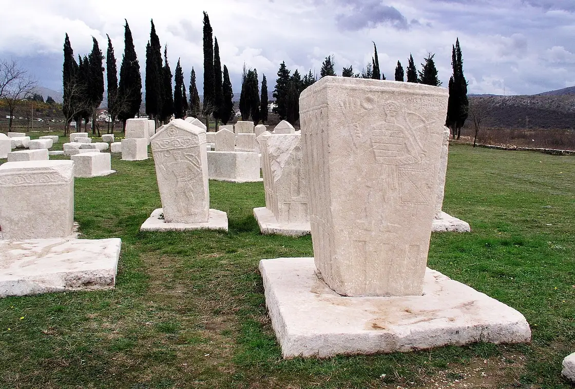 A row of tall chest stecci on pedestals at Radimlja, the nearest carved with a standing figure, cypress trees and karst hills behind