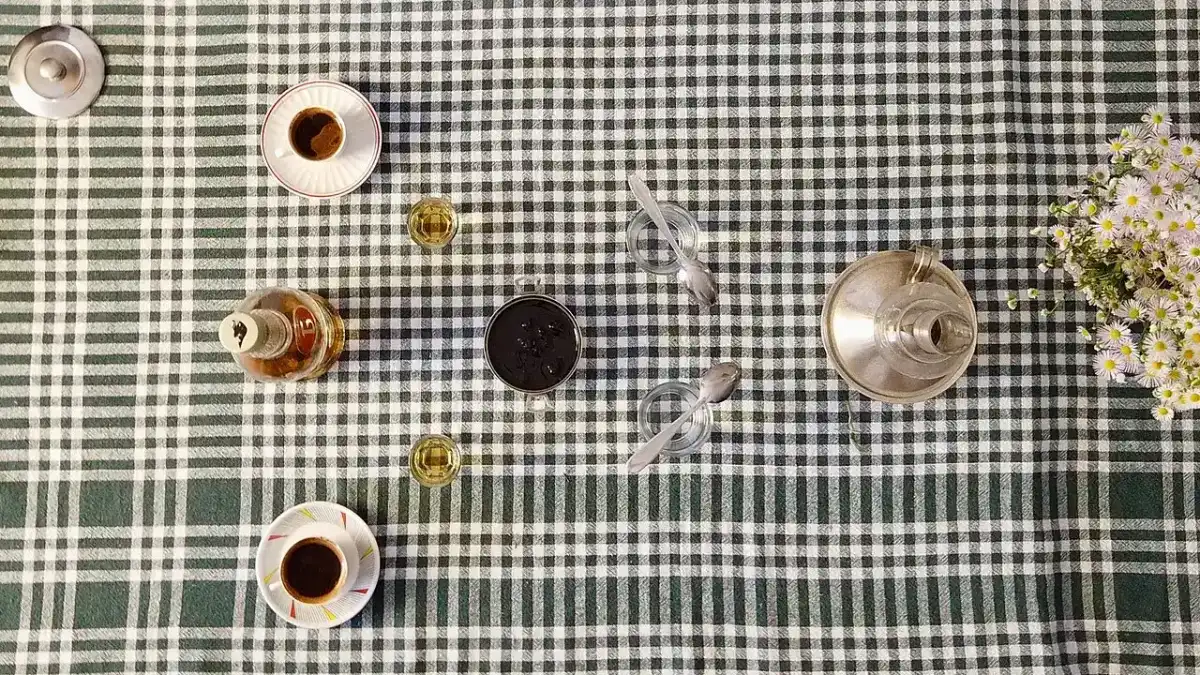 An overhead view of Bosnian coffee, small glasses of rakija and a jar of slatko fruit preserve laid out on a checked tablecloth