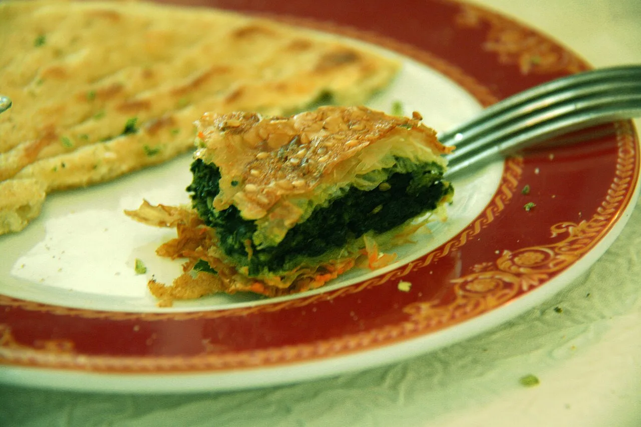 A cut slice of flaky spinach filo pastry on a decorative plate next to a whole piece of the pie, with a fork resting beside it