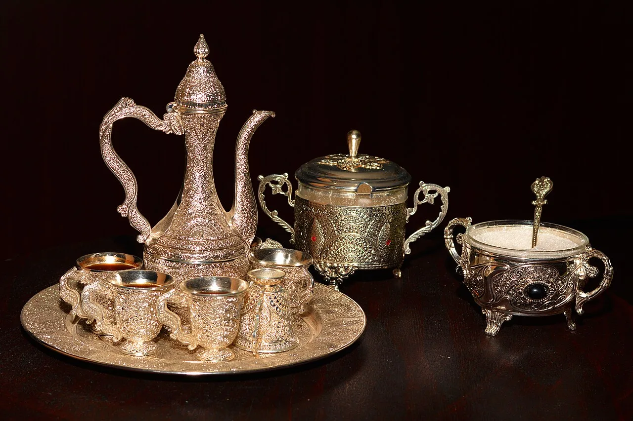 An ornate engraved coffee service set with a tall pot, small cups on a round tray and two lidded sugar bowls